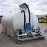 An 8000L Civmaster Premium Hydraulic Water Cart by National Water Carts, featuring attached hoses, pipes, and valves on a metal frame, stands outdoors on concrete with buildings, cars, and trees visible beneath a cloudy sky.