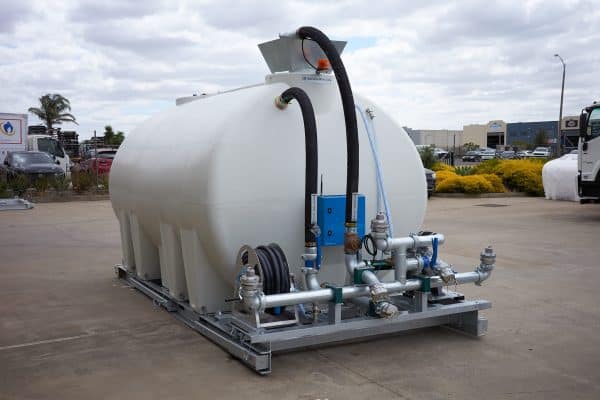 An 8000L Civmaster Premium Hydraulic Water Cart by National Water Carts, featuring attached hoses, pipes, and valves on a metal frame, stands outdoors on concrete with buildings, cars, and trees visible beneath a cloudy sky.