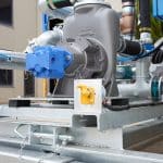 Close-up of the 8000L Civmaster Premium Hydraulic Water Cart showing metal pipes, a blue valve, and a gray pump; a small yellow control switch is mounted on the frame. The buildings structure is visible in the background.
