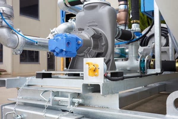 Close-up of the 8000L Civmaster Premium Hydraulic Water Cart showing metal pipes, a blue valve, and a gray pump; a small yellow control switch is mounted on the frame. The buildings structure is visible in the background.