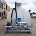 The 8000L Civmaster Premium Hydraulic Water Cart by National Water Carts, featuring metal pipes, hoses, and valves on a frame, is positioned outdoors on concrete near commercial buildings and parked vehicles.