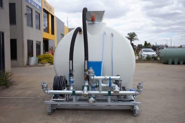 The 8000L Civmaster Premium Hydraulic Water Cart by National Water Carts, featuring metal pipes, hoses, and valves on a frame, is positioned outdoors on concrete near commercial buildings and parked vehicles.