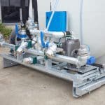 The 8000L Civmaster Premium Hydraulic Water Cart features pipes, valves, and gauges on a metal frame, positioned outdoors near a building and greenery.