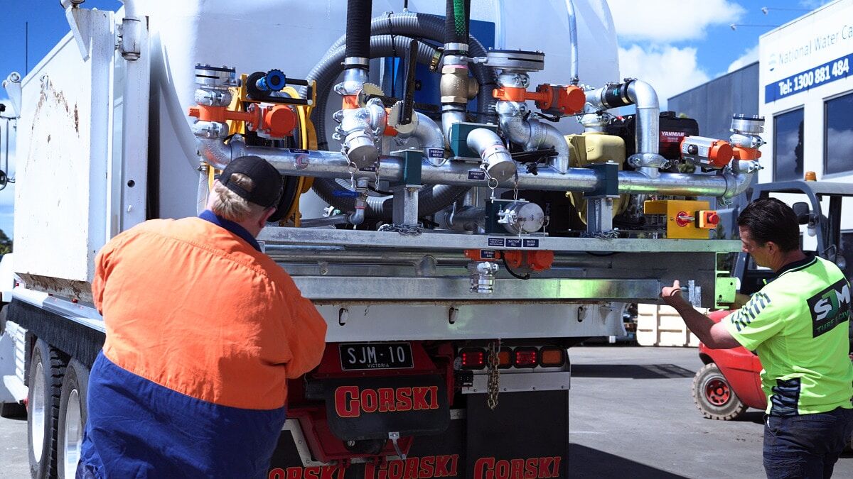 Two workers in high-visibility PPE inspect the pipe manifold on the rear of a CivMaster Premium slip-on water cart at an outdoor worksite.