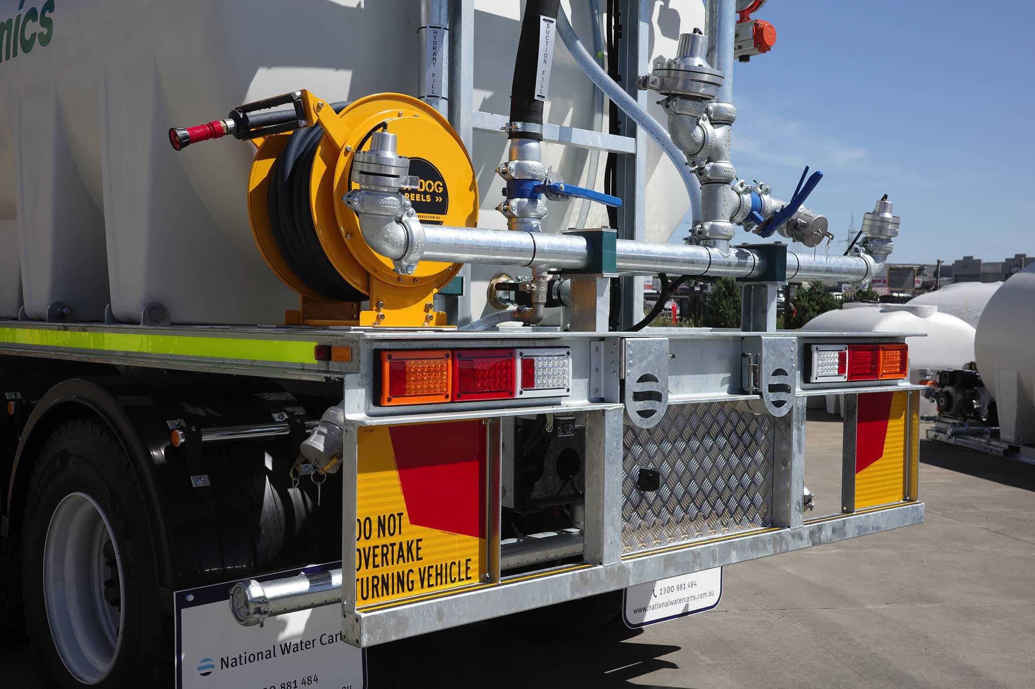 A rear view of a CivMaster EXM water cart showing the spray system manifold with hoses, pipes, valves, and rear safety equipment.