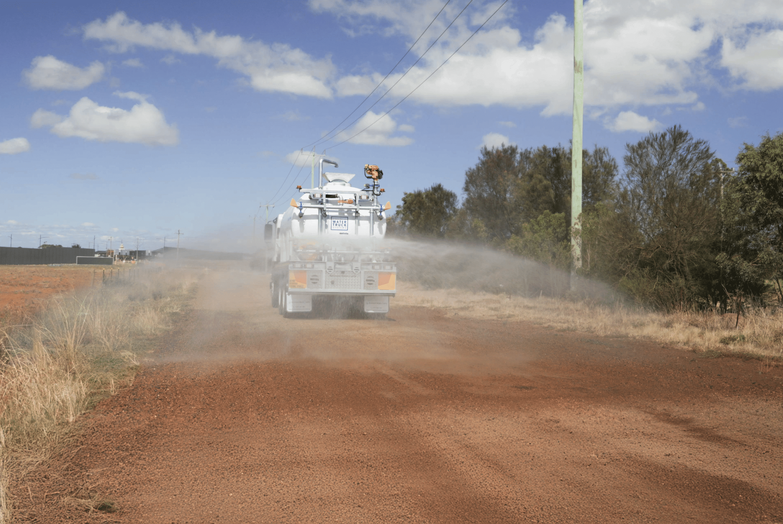 A National Water Carts 13,000-litre FVZ water truck uses a rear centre spray head to suppress dust on a rural dirt road.