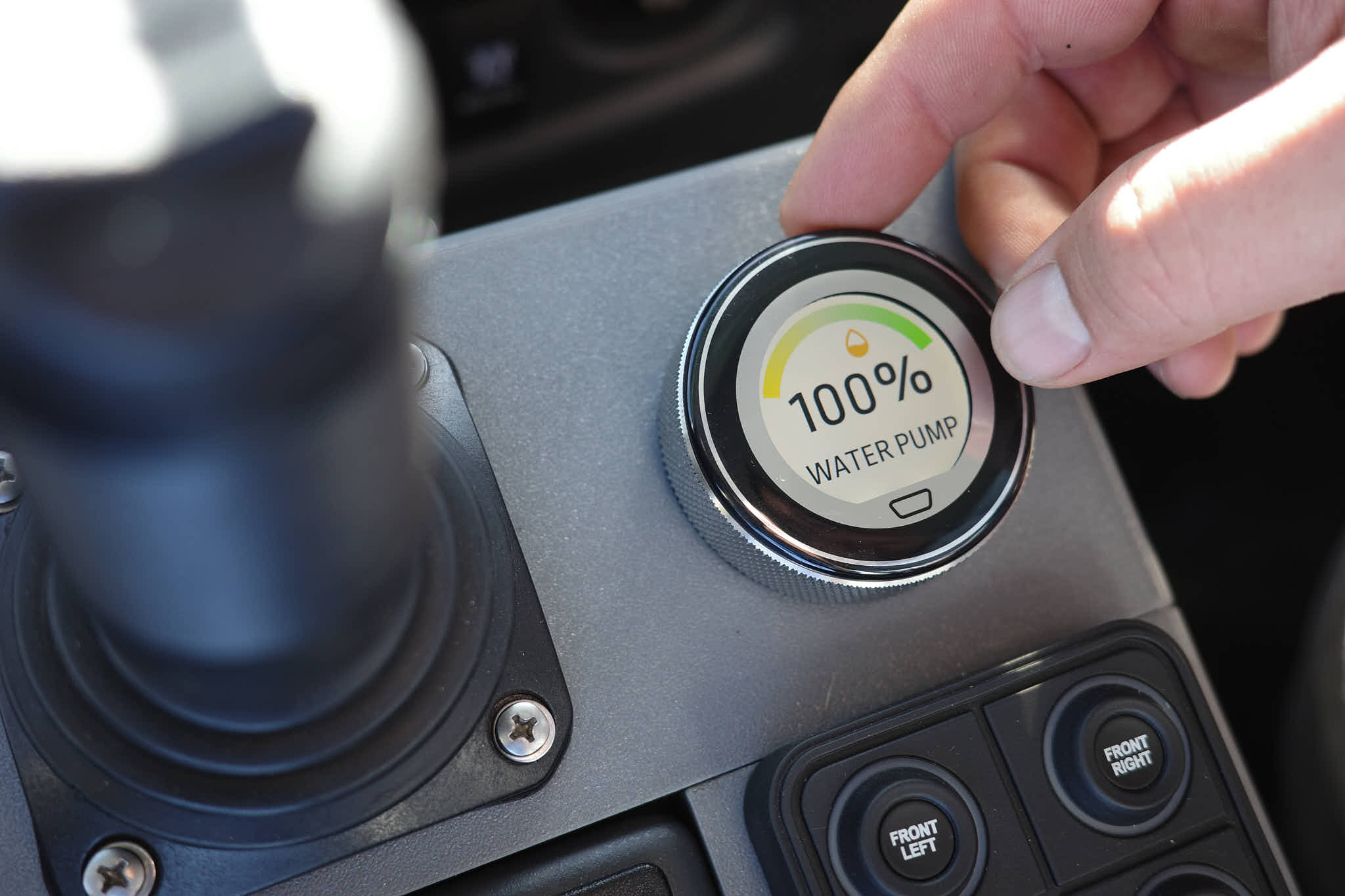 A hand adjusts the CANbus control panel on a water truck, showing a digital water pump readout at 100 percent output.