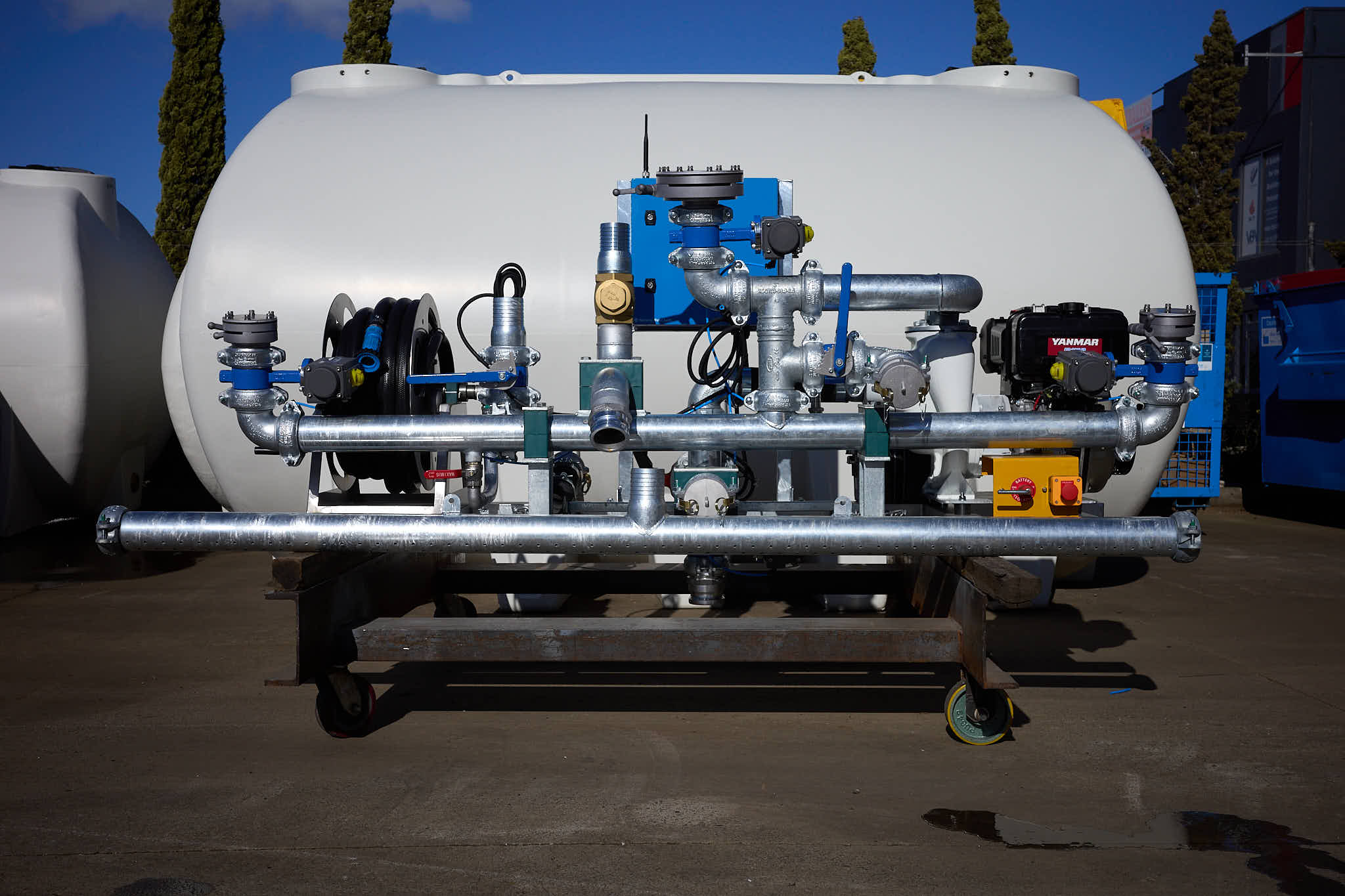 A large industrial tank with metal pipes, valves, gauges, and machinery—part of a diesel spray system—mounted on a platform and standing outdoors on a concrete surface under a blue sky.