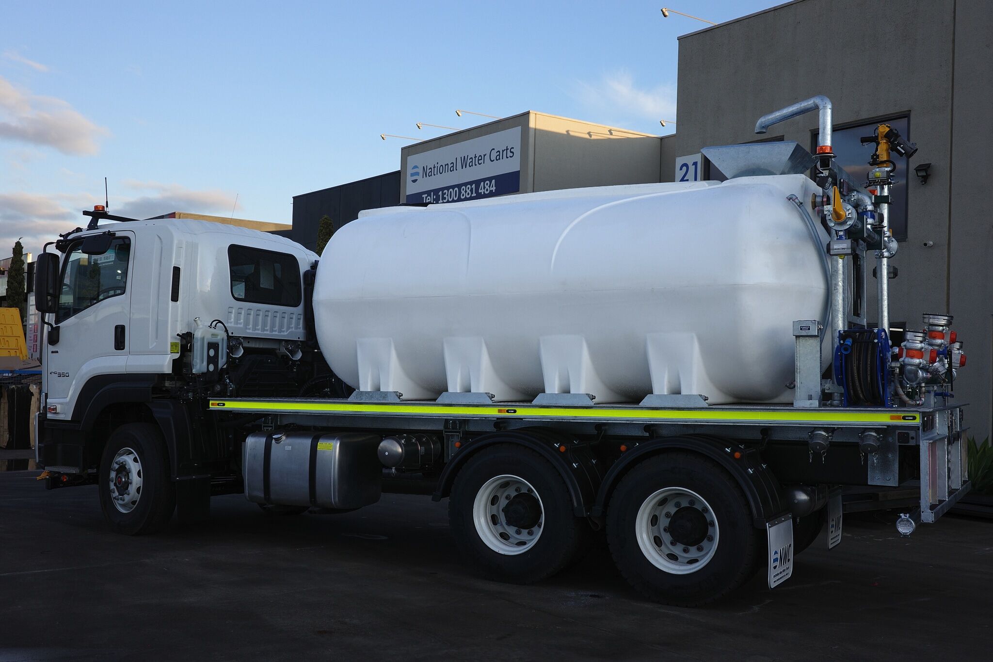 A CANbus Hydraulic Water Truck with a large cylindrical tank is parked outside a commercial building. The tank, supplied by National Water Carts, is mounted on the truck bed with pipes and valves attached at the rear.