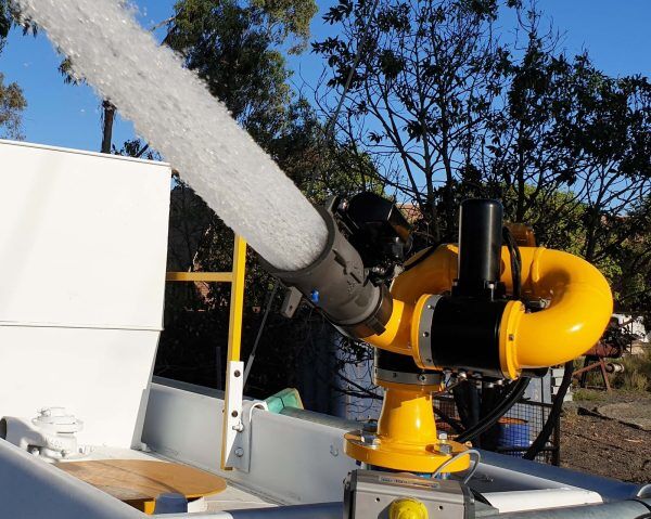 The E360XD Extreme Duty Electric Water Cannon by National Water Carts sprays a strong stream of water, mounted on a white vehicle with trees and outdoor equipment seen in the background.