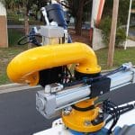 A close-up of the E360XD Extreme Duty Electric Water Cannon, featuring a bright yellow industrial robotic arm with sensors and wires, is mounted outdoors on a platform near a street and buildings by National Water Carts.