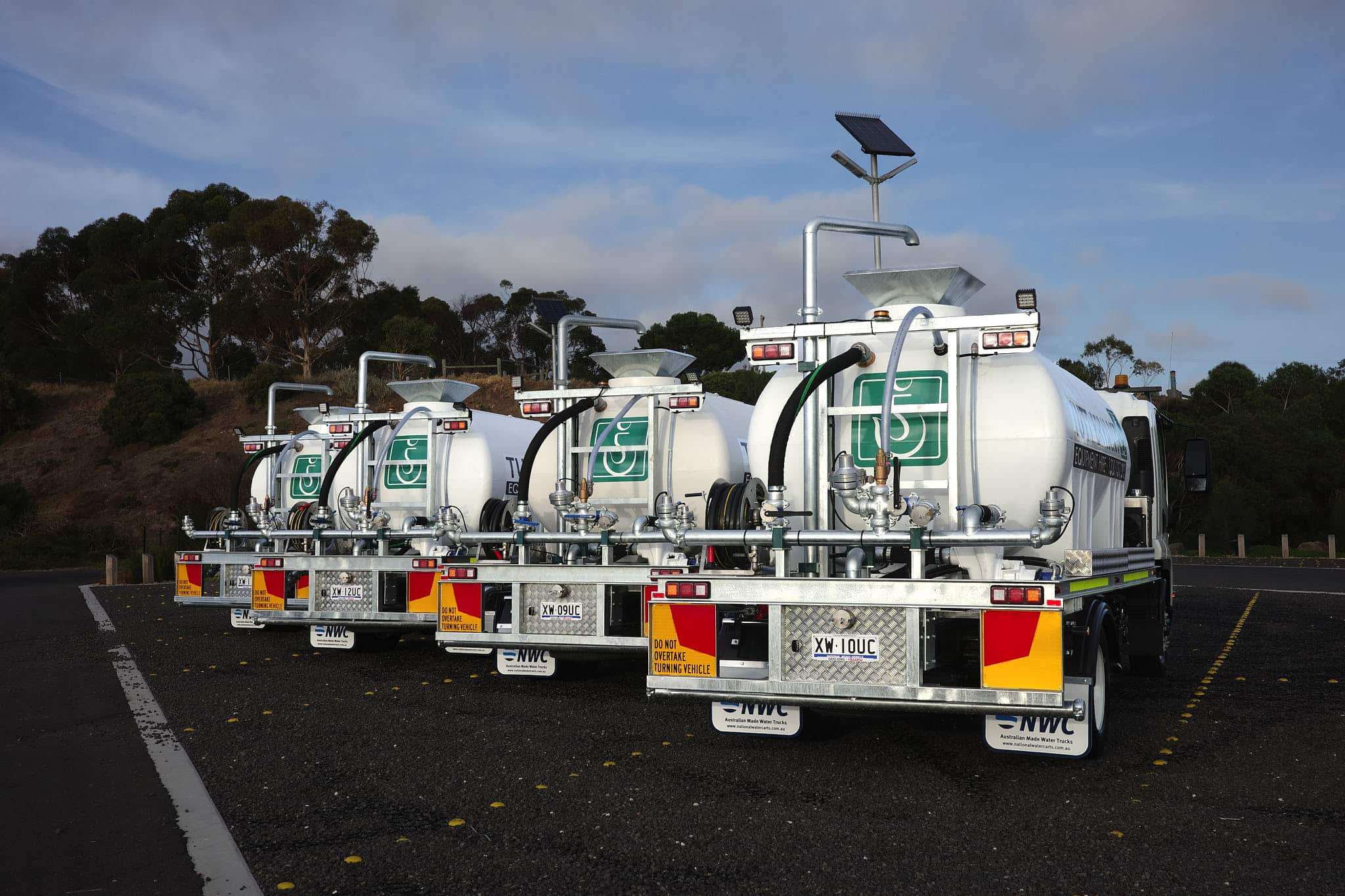 Four white water tank trucks with green logos are parked in a row on asphalt, each equipped with hoses, metal fittings, and solar panels, with trees and a clear sky in the background.